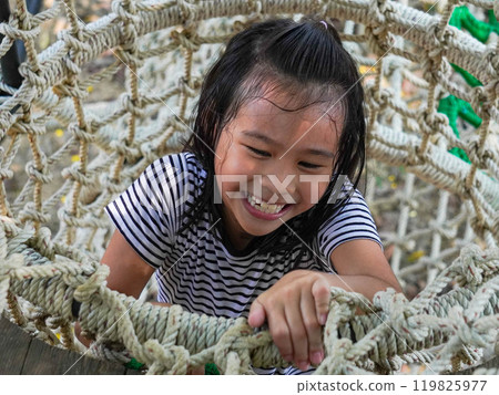 Children play outdoors on a wooden playground in a park. Happy girl playing on a rope net bridge in playground during summer. Children play outdoors on a wooden playground in a park. Happy girl playing on a rope net bridge in playground during summer. 119825977