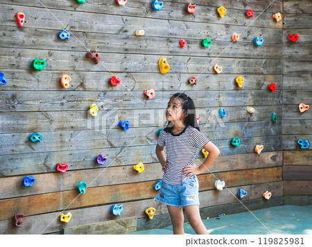 Children play outdoors on a wooden playground in a park. Happy girl playing on wooden climbing wall in playground during summer. Children play outdoors on a wooden playground in a park. Happy girl playing on wooden climbing wall in playground during summer. 119825981
