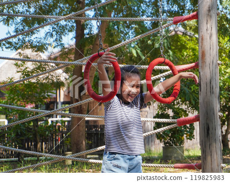 Children play outdoors on a wooden playground in a park. Happy girl playing on climbing swings in playground during summer. Children play outdoors on a wooden playground in a park. Happy girl playing on climbing swings in playground during summer. 119825983