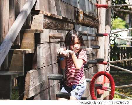 Children play outdoors on a wooden playground in a park. Happy girl playing on wooden climbing wall in playground during summer. Children play outdoors on a wooden playground in a park. Happy girl playing on wooden climbing wall in playground during summer. 119826070