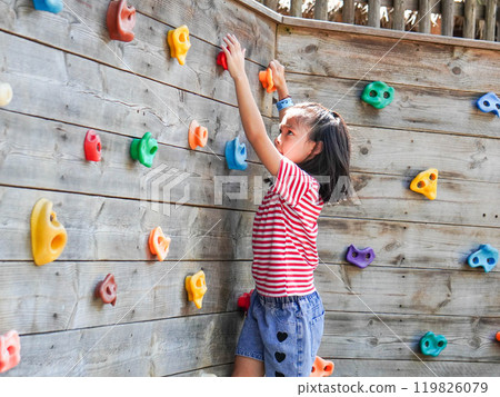 Children play outdoors on a wooden playground in a park. Happy girl playing on wooden climbing wall in playground during summer. 119826079