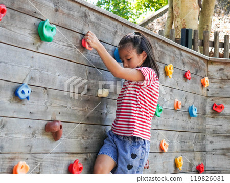 Children play outdoors on a wooden playground in a park. Happy girl playing on wooden climbing wall in playground during summer. 119826081