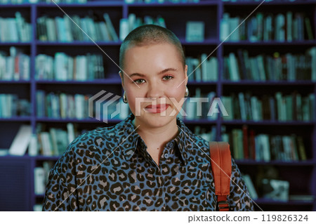 Portrait of smiling woman wearing leopard-print shirt with shelves full of books in background, bald hairstyle, holding a bag with strap over shoulder in the library setting 119826324