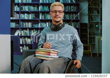 Portrait of man sitting in wheelchair, holding stack of books and wearing glasses. Bookshelves and diverse collection of books are visible in background Portrait of man sitting in wheelchair, holding stack of books and wearing glasses. Bookshelves and diverse collection of books are visible in background 119826337