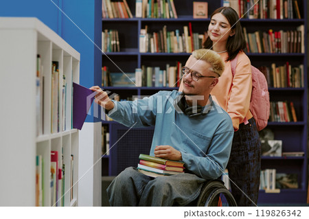 Two students exploring books in library, one in wheelchair perusing a new book while other standing and observing the process 119826342