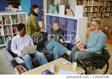 Group of diverse students studying in library, engaging in discussion with textbooks and laptops around. Bright, spacious study area surrounded by bookshelves 119826386