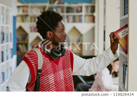 Young African American person selecting data analysis book in library, wearing red vest and headphones around neck. Bookshelf and other library users in background Young African American person selecting data analysis book in library, wearing red vest and headphones around neck. Bookshelf and other library users in background 119826434