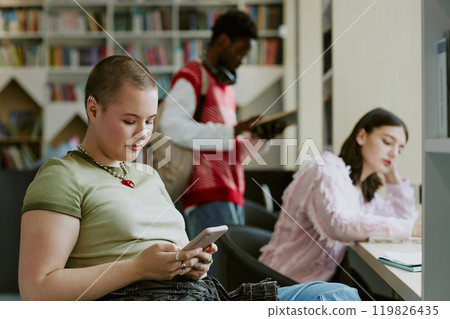 Students studying in a library setting with bookshelves in background, using smartphones and reading material. Scene captures focus and concentration while engaging with digital and printed resources 119826435