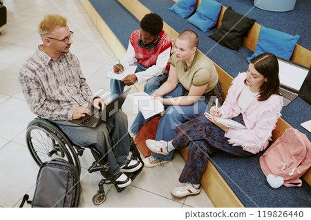 Group of diverse university students engaging in studying, sitting on campus steps, with notes and laptops, involving student in wheelchair, promoting collaborative learning atmosphere Group of diverse university students engaging in studying, sitting on campus steps, with notes and laptops, involving student in wheelchair, promoting collaborative learning atmosphere 119826440