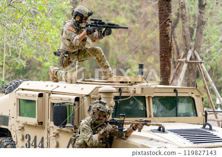 Two militaries or soldiers stay near humvee car with on on roof and the other one in area of car hood also point gun to right side during outdoor practice. 119827143