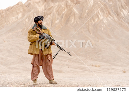 The man with costume look like terrorist hold gun and stand in front of mountain and look forward. 119827275