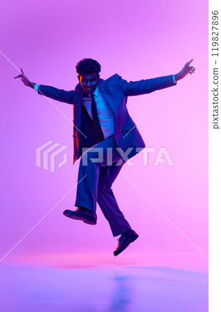 Full-length portrait of happy young Indian man, businessman in formal wear dancing, jumping in joy against purple studio background in neon light 119827896