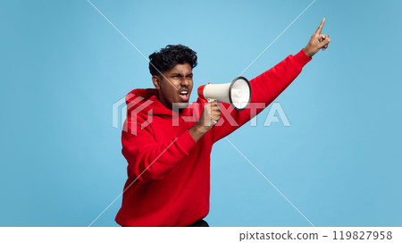 Emotional young Indian man shouting in megaphone wand lively gesturing against blue studio background. Announcement, news, propaganda Emotional young Indian man shouting in megaphone wand lively gesturing against blue studio background. Announcement, news, propaganda 119827958
