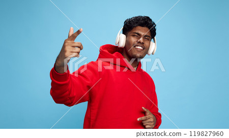 Portrait of young Indian man in red oversized hoodie, listening to energetic music in headphones and singing against blue studio background 119827960