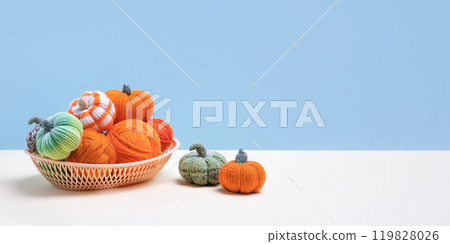Knitted orange and green pumpkins in basket with knitting balls on blue background with space for text, knitting hobby, autumn composition. Knitted orange and green pumpkins in basket with knitting balls on blue background with space for text, knitting hobby, autumn composition. 119828026
