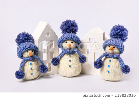 Knitted snowman in blue hat and scarf with white houses on gray background. Winter fairy tale story with your own hands, hobby. Merry Christmas and New Year 2025 greeting card 119828030
