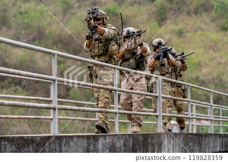 Group of militaries or soldiers with battle suit hold and point gun and walk in the row on bridge during outdoor practice. 119828359