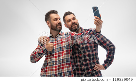 Two young guys, twin brothers dressed matching checked shirts and jeans, hugs and taking selfie against white studio background. 119829049