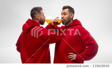 Two identically dressed young guys, twin brothers, drink craft beer from glass goblets against white studio background. 119829050