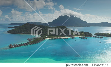 Luxurious overwater bungalows line the shores of tropical island paradise, lush green mountains rising in background, creating an idyllic vacation destination. Remote wild nature travel. Aerial view Luxurious overwater bungalows line the shores of tropical island paradise, lush green mountains rising in background, creating an idyllic vacation destination. Remote wild nature travel. Aerial view 119829334