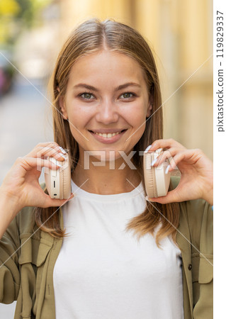 Happy Caucasian young woman listening to music taking off headphones looking at camera on street 119829337