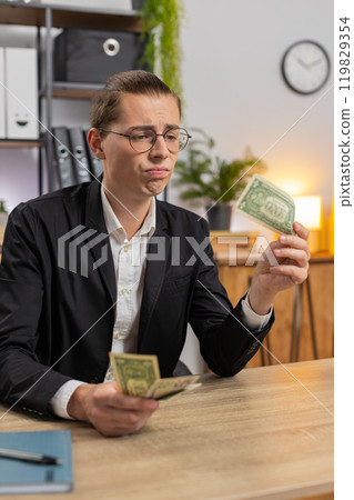 Upset young businessman freelancer counting dollar cash insufficient amount of money at office desk Upset young businessman freelancer counting dollar cash insufficient amount of money at office desk 119829354