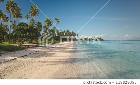 Paradise island beach with palm trees and turquoise blue water gently laps white sand shore in sunny day. Tourists enjoying tropical paradise. Remote wild nature, exotic summer travel background Paradise island beach with palm trees and turquoise blue water gently laps white sand shore in sunny day. Tourists enjoying tropical paradise. Remote wild nature, exotic summer travel background 119829355