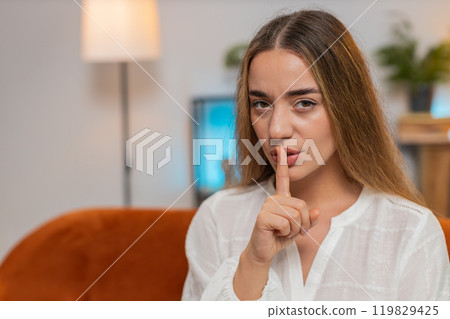 Portrait of young woman on sofa presses index finger to lips makes silence hush gesture sign at home 119829425