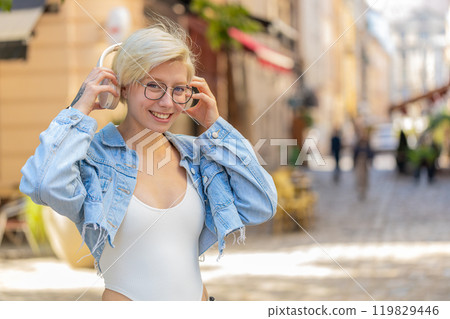 Portrait of smiling young woman in glasses taking off wireless headphones while looking at camera Portrait of smiling young woman in glasses taking off wireless headphones while looking at camera 119829446