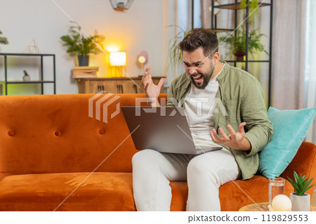 Upset young Caucasian man freelancer using laptop engages in heated conversations sitting on sofa Upset young Caucasian man freelancer using laptop engages in heated conversations sitting on sofa 119829553