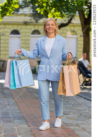 Mature businesswoman in suit carrying shopping bags while walking on city street after purchases Mature businesswoman in suit carrying shopping bags while walking on city street after purchases 119829555