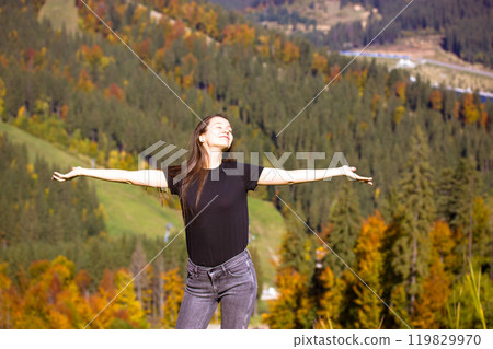 Happy young white woman, girl in jeans, black T-shirt standing with arms outstretched against autumnal landscape high in the mountains on an autumn day. Travel in autumn, fall season. Bucovel Ukraine. Happy young white woman, girl in jeans, black T-shirt standing with arms outstretched against autumnal landscape high in the mountains on an autumn day. Travel in autumn, fall season. Bucovel Ukraine. 119829970