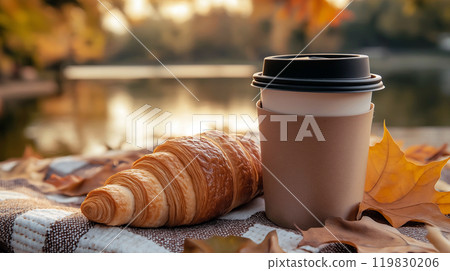 Coffee or tea cup and croissant on picnic plaid near the lake in autumn park. Cozy autumn picnic set up outdoors in the morning, breakfast in nature 119830206