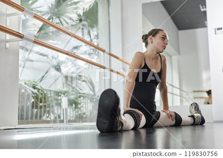 Side view of young thoughtful girl dressed in elegant black leotard sitting on floor perfecting splits while stretching legs before ballet class in studio with huge windows, copy space Side view of young thoughtful girl dressed in elegant black leotard sitting on floor perfecting splits while stretching legs before ballet class in studio with huge windows, copy space 119830756