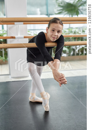 Vertical shot of young ballerina in pointe shoes cheerfully smiling posing for camera while leaning on barre in dance studio 119830758