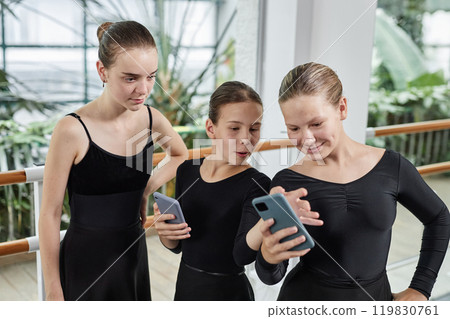 Three young female ballet dancers looking at phone screen watching funny video while relaxing after dance practicing in studio 119830761