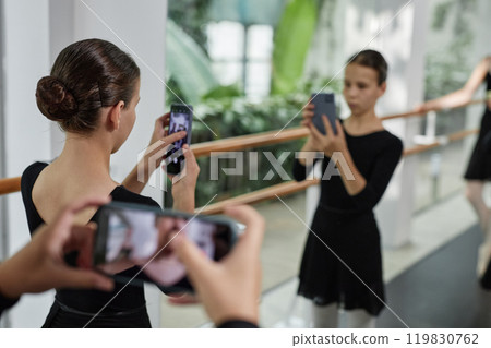 Rear view of little girl with hair styled in bun taking photo in mirror during ballet class in dance studio, copy space 119830762