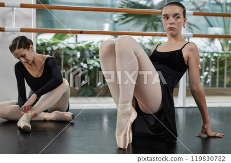 Elegant young ballet dancer standing on tiptoes checking pointe shoes dressing while preparing for dance class sitting on floor in studio, copy space Elegant young ballet dancer standing on tiptoes checking pointe shoes dressing while preparing for dance class sitting on floor in studio, copy space 119830782