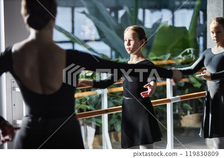 Concentrated young girl practicing ballet positions standing at barre in sunlight during group class in dance studio decorated with green plants Concentrated young girl practicing ballet positions standing at barre in sunlight during group class in dance studio decorated with green plants 119830809