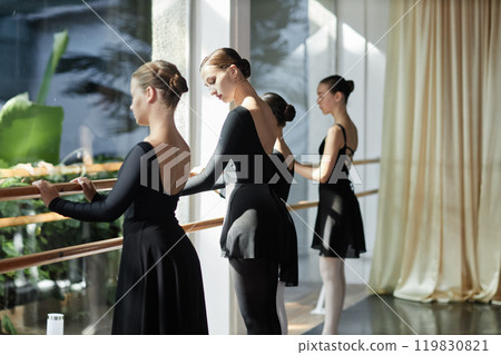 Medium shot of young woman in elegant black leotard teaching children dance positions standing at barre during ballet class in sunlit studio, copy space Medium shot of young woman in elegant black leotard teaching children dance positions standing at barre during ballet class in sunlit studio, copy space 119830821