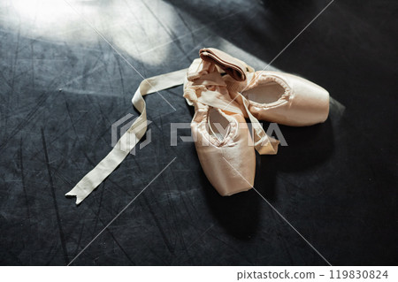 Background shot of beige satin pointe shoes decorated with elegant ribbons lying in sunlight on dark floor in dance studio , copy space 119830824