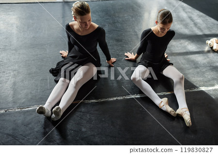 High angle shot of two little female ballet dancers in black leotards sitting on floor stretching leg muscles, while preparing for group class in dance studio High angle shot of two little female ballet dancers in black leotards sitting on floor stretching leg muscles, while preparing for group class in dance studio 119830827