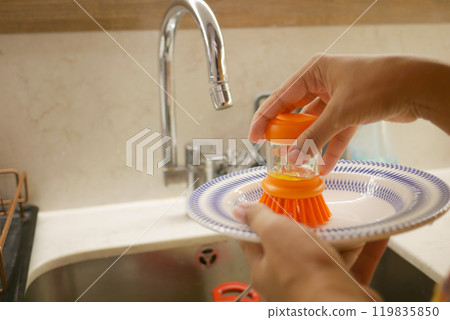 Hands scrub a plate with an orange brush in a sink, emphasizing cleanliness and hygiene Hands scrub a plate with an orange brush in a sink, emphasizing cleanliness and hygiene 119835850