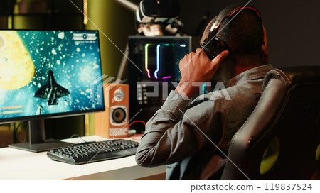 Man playing SF shooter videogame on computer monitor, talking into headset with friends. Gamer using headphones while enjoying game with spaceship flying to chat with Esports team, camera A 119837524
