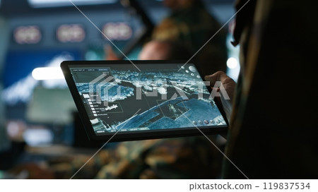 Military agent working in control room, using software on tablet, analyzing aerial reconnaissance data. Close up of intelligence corps expert controlling drones with portable device, camera A Military agent working in control room, using software on tablet, analyzing aerial reconnaissance data. Close up of intelligence corps expert controlling drones with portable device, camera A 119837534