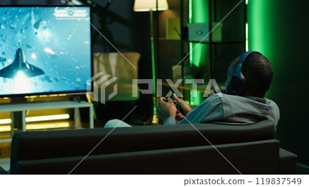 Man playing entertaining spaceship flying videogame on large smart TV, wearing headphones. Player enjoying SF arcade game on gaming console, piloting spacecraft in galaxy using gamepad, camera A Man playing entertaining spaceship flying videogame on large smart TV, wearing headphones. Player enjoying SF arcade game on gaming console, piloting spacecraft in galaxy using gamepad, camera A 119837549