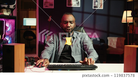 Man in neon lit apartment playing intense videogames on gaming PC at computer desk. Gamer competing against enemies during internet multiplayer competition, unwinding at home, camera B 119837578
