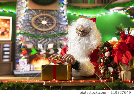 Saint Nicholas prepares a festive gift box by the fire. The North Pole workshop is alive with holiday decorations and the magic of giving, as Santa packs presents for children all over the world. Saint Nicholas prepares a festive gift box by the fire. The North Pole workshop is alive with holiday decorations and the magic of giving, as Santa packs presents for children all over the world. 119837649