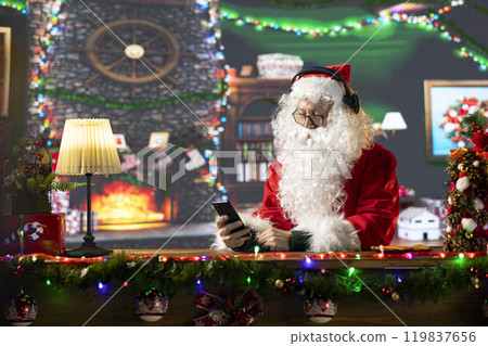 Happy old Santa Claus listening to Christmas carols and festive music on his smartphone, white bearded man in suit spreading the holiday cheer in log cabin. Saint Nicholas uses headphones. Happy old Santa Claus listening to Christmas carols and festive music on his smartphone, white bearded man in suit spreading the holiday cheer in log cabin. Saint Nicholas uses headphones. 119837656