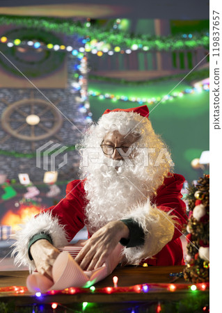 Santa Claus decorates a gift box in his festive workshop preparing wrapped presents and ornaments to fill the holiday season with magic. Christmas delivery from the North Pole, tradition and joy. Santa Claus decorates a gift box in his festive workshop preparing wrapped presents and ornaments to fill the holiday season with magic. Christmas delivery from the North Pole, tradition and joy. 119837657